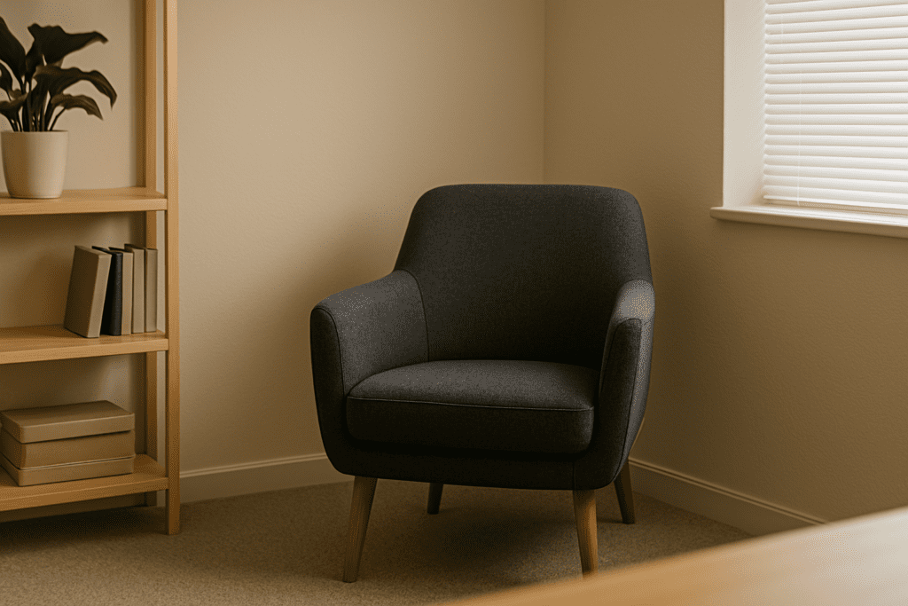 A clean, modern therapy room corner with a gray armchair and wooden shelving under natural daylight, evoking a peaceful atmosphere.