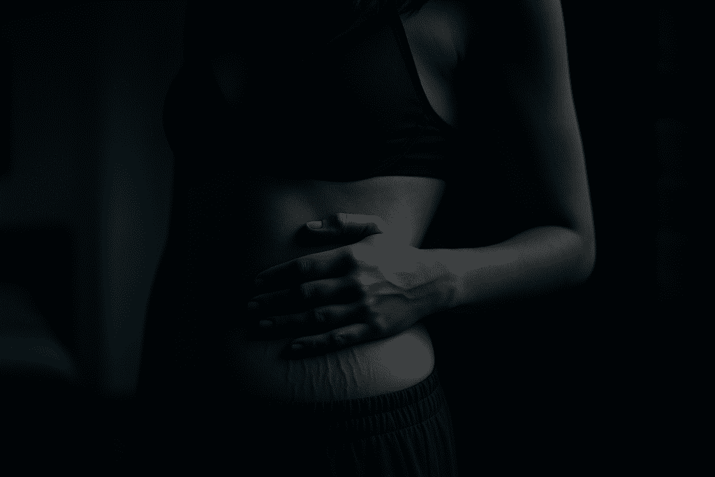In a low-lit indoor scene, a woman gently places her hand over stretch marks on her bare torso, illuminated by a cool, directional light. The dramatic shadows and muted tones evoke an introspective mood, underscoring the emotional and physical journey of managing EDS-related skin conditions.